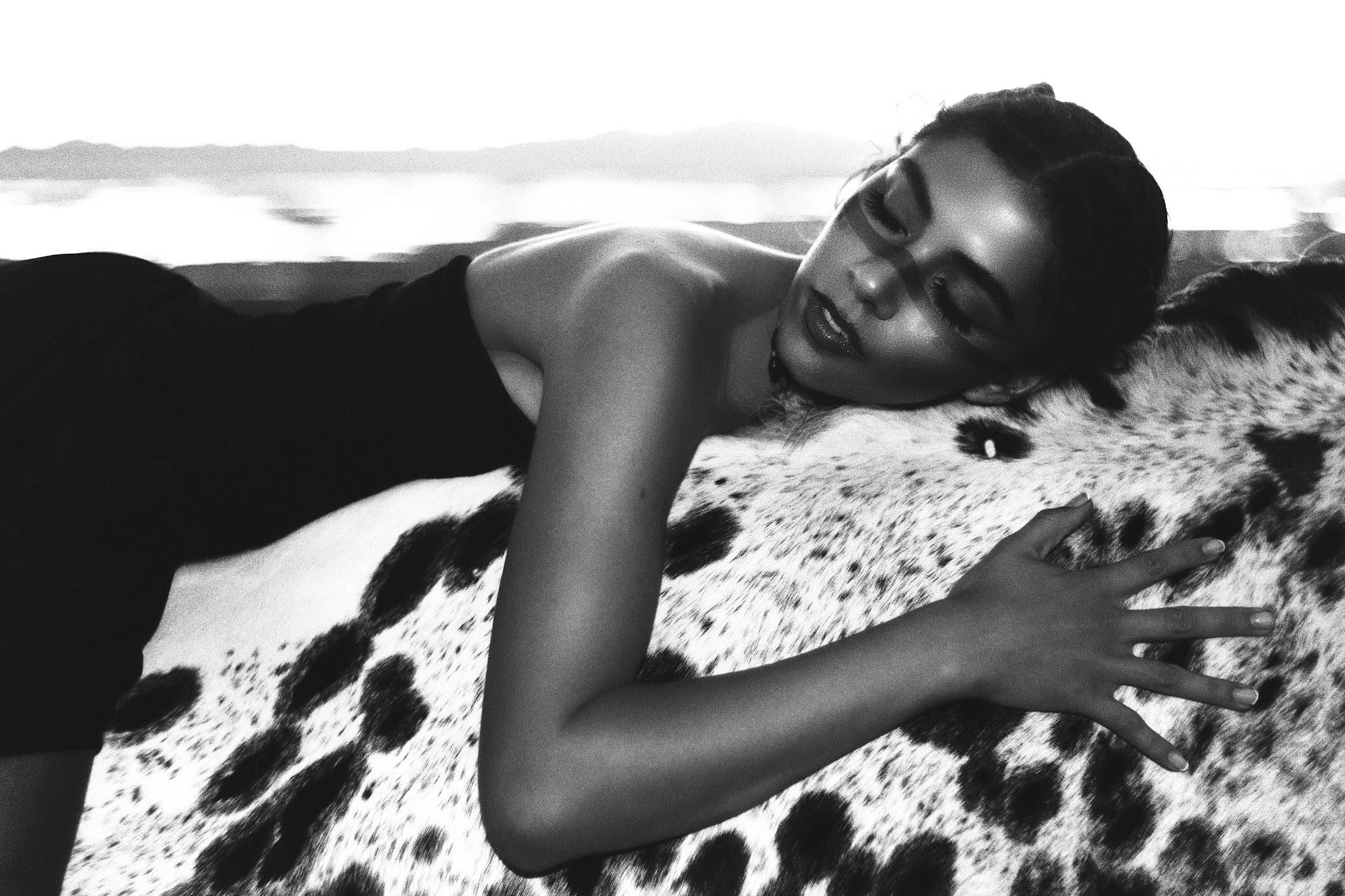 A woman resting against a spotted horse coat, mountains in the distance — black and white