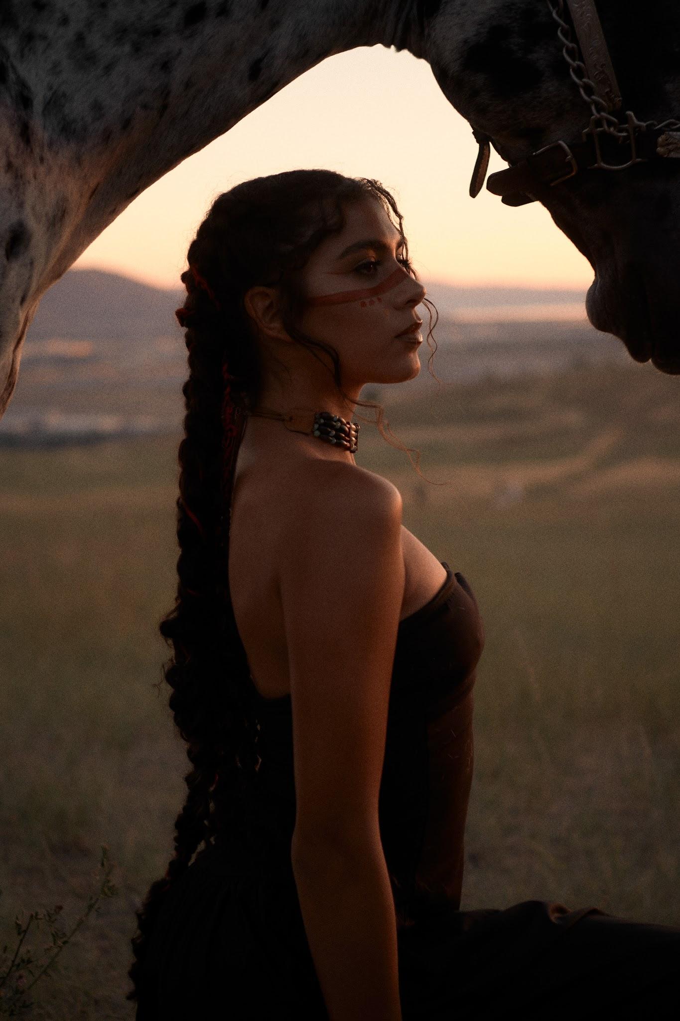 A young Diné woman with traditional face paint beside her horse at sunset